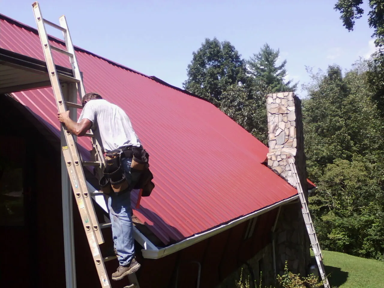 Roofer on a ladder beside a red metal roof and stone chimney — Soffit & Fascia Repair by Roofing Brothers in Pierce